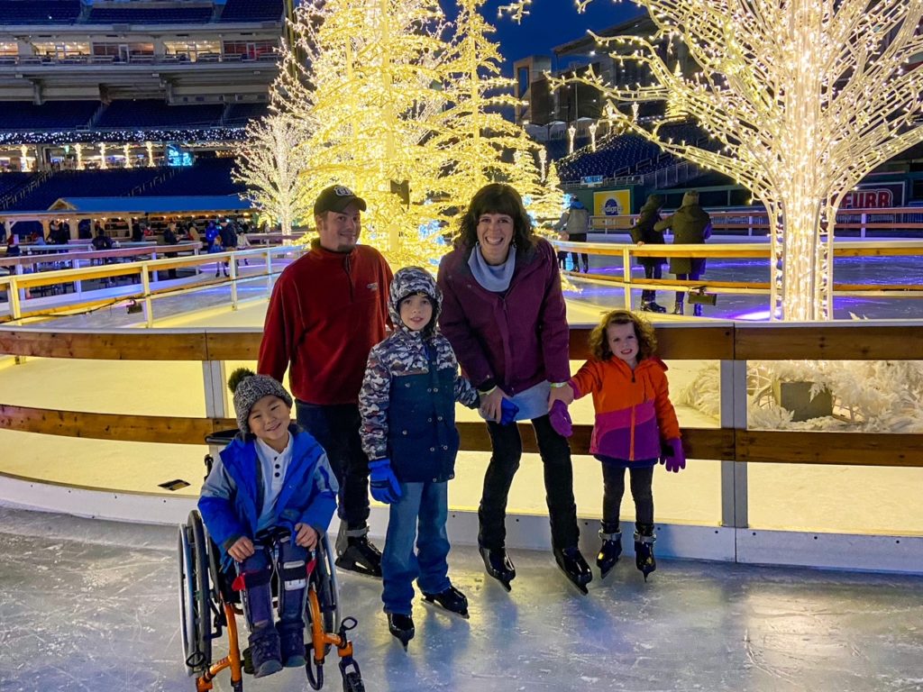 Boy in wheelchair smiles on accessible ice rink with his family in ice skates, surrounded by christmas lights