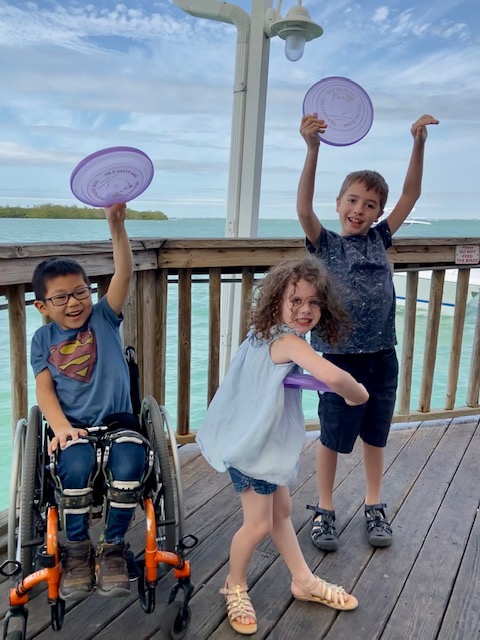 Little boy in orange wheelchair with little sister and bog brother  smiling and pretending to throw purple frisbees. All three in blue shirts, with the ocean behind them. 