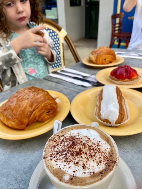Cappuccino, croissants, and French pastries across a gray table. Little girl in background