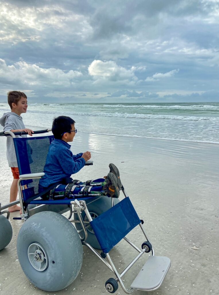 boy in beach wheelchair with big brother behind him. both looking at the waves.