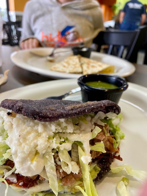 Dark blue hard taco shell overflowing with soft cheese, lettuce, and other hard to distinguish foods.