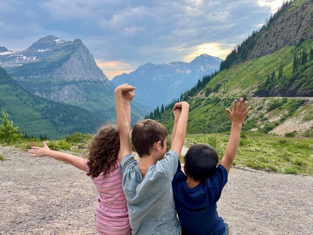 three kids hold hands up in the air with mountain view behind them