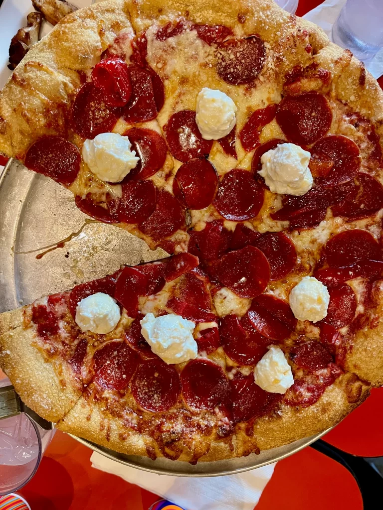 Pizza with one slice out of it. Pizza is topped with pepperoni, peppercinis, and dollops of ricotta