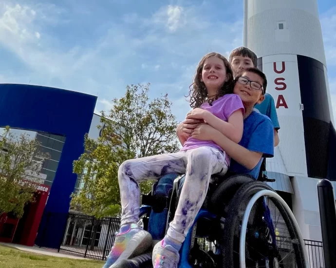 boy in wheelchair in front of massive model rocket. Little sister in his lap, big brother standing behind.