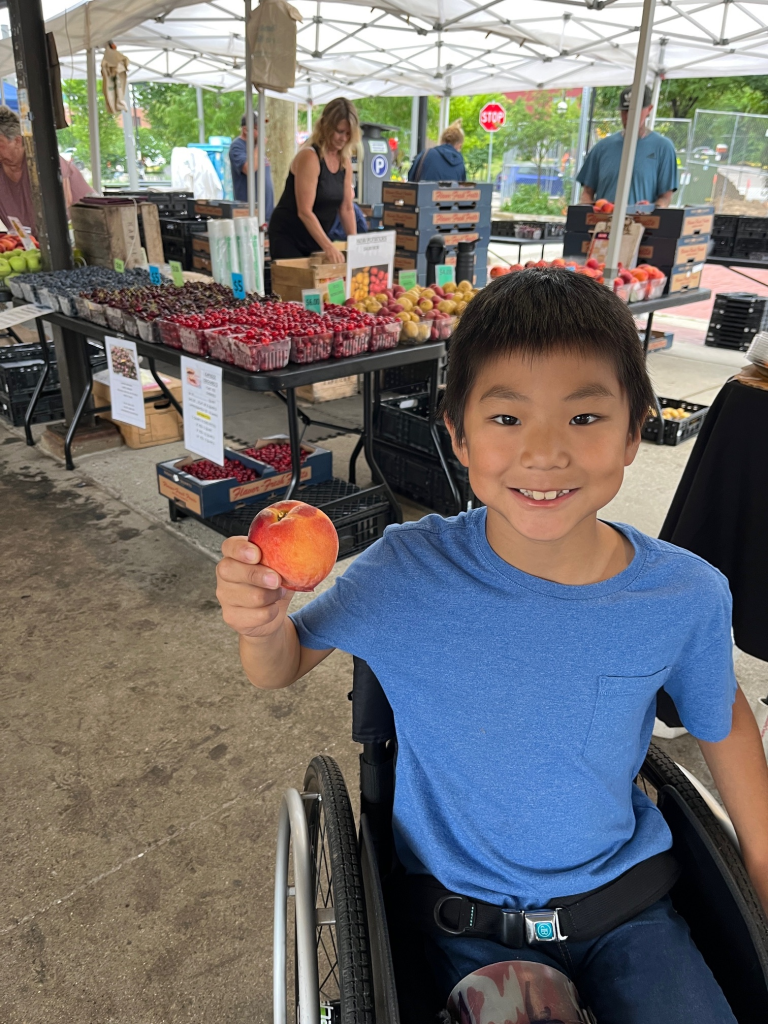little boy in wheelchair holding up a peach with a fruit stand behind him