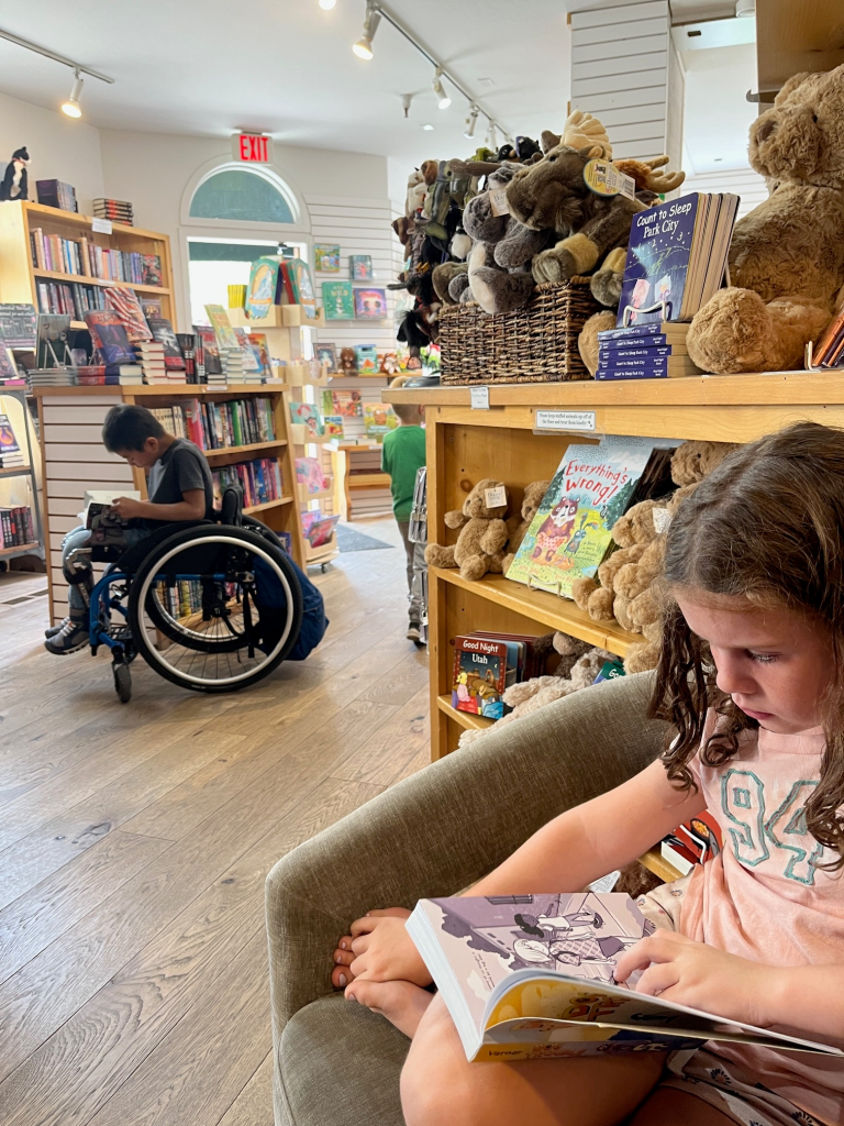 boy in wheelchair absorbed in a book in the bookstore aisle. Little sister in foreground reading in armchair.