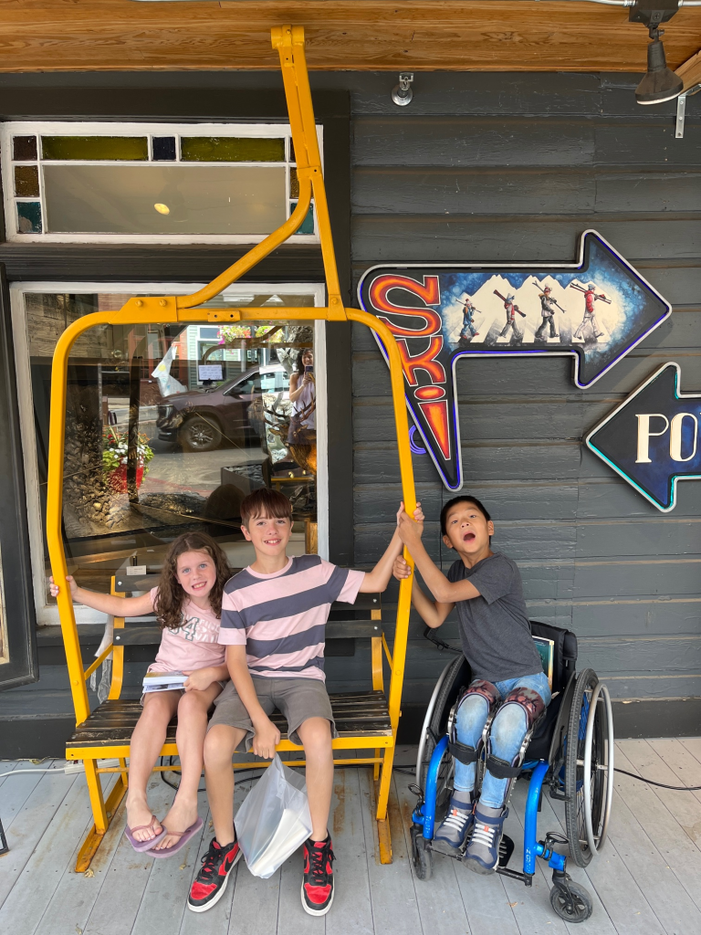preteen boy and little sister on ski lift bench on the sidewalk. Boy in wheelchair hangs on the end pretending to be screaming through the air. "Ski" sign in the background.