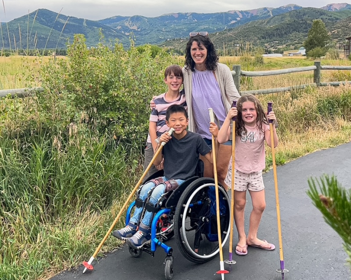 boy in wheelchair with brother, sister, and mom on paved trail in front of mountains