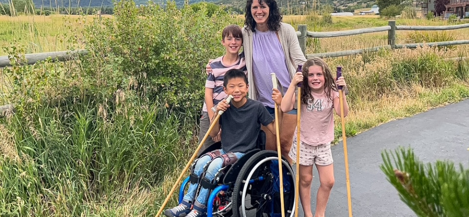 boy in wheelchair with brother, sister, and mom on paved trail in front of mountains