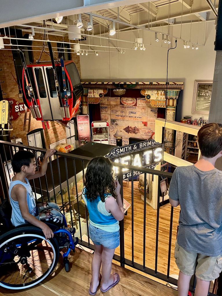 gondola hangs from the ceiling above an old fashioned store front. Kids in the foreground are looking at the scene over the balcony.