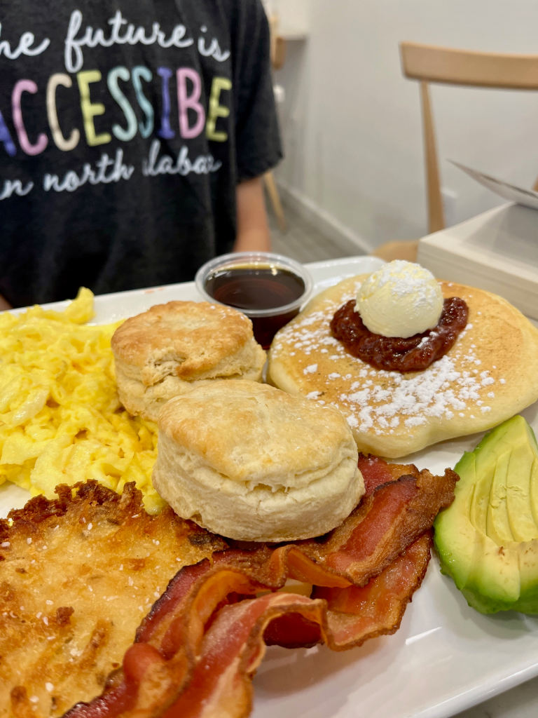 two biscuits, scrambled eggs, pancake with red jelly on top and syrup beside, sliced avocado, two slices of bacon, and a hash brown cake