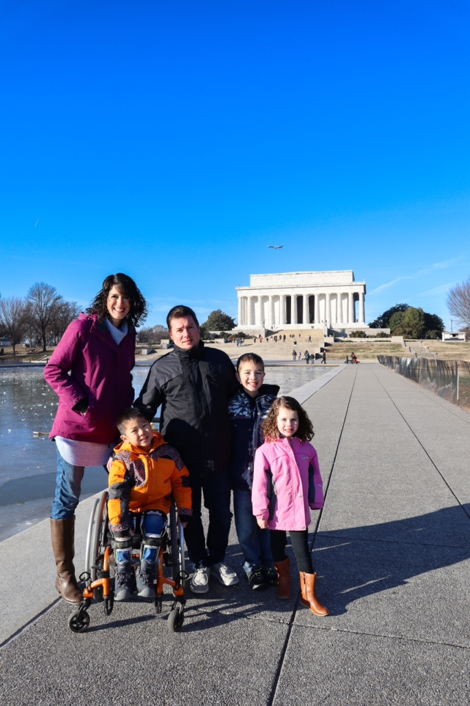 Family in winter coats smiles for camera beside long pool of water leading to the columns of the Lincoln Memorial