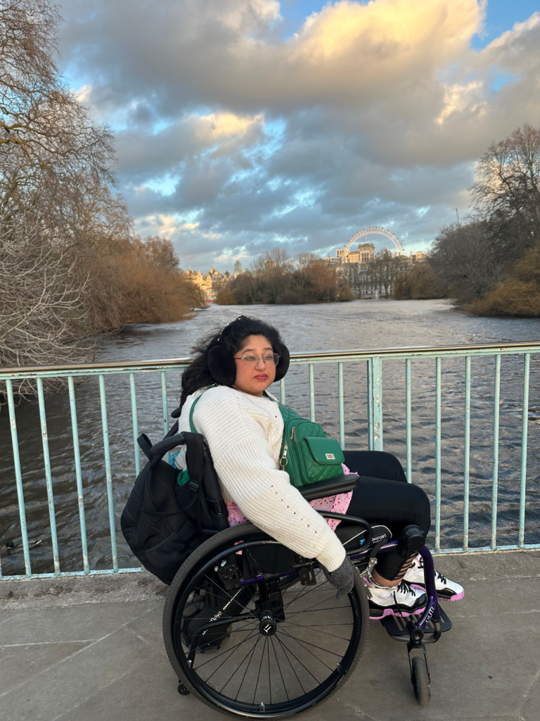 Hasitha in her chair on a bridge with the large ferris wheel in the background. She's wearing ear muffs and gloves and looks cold
