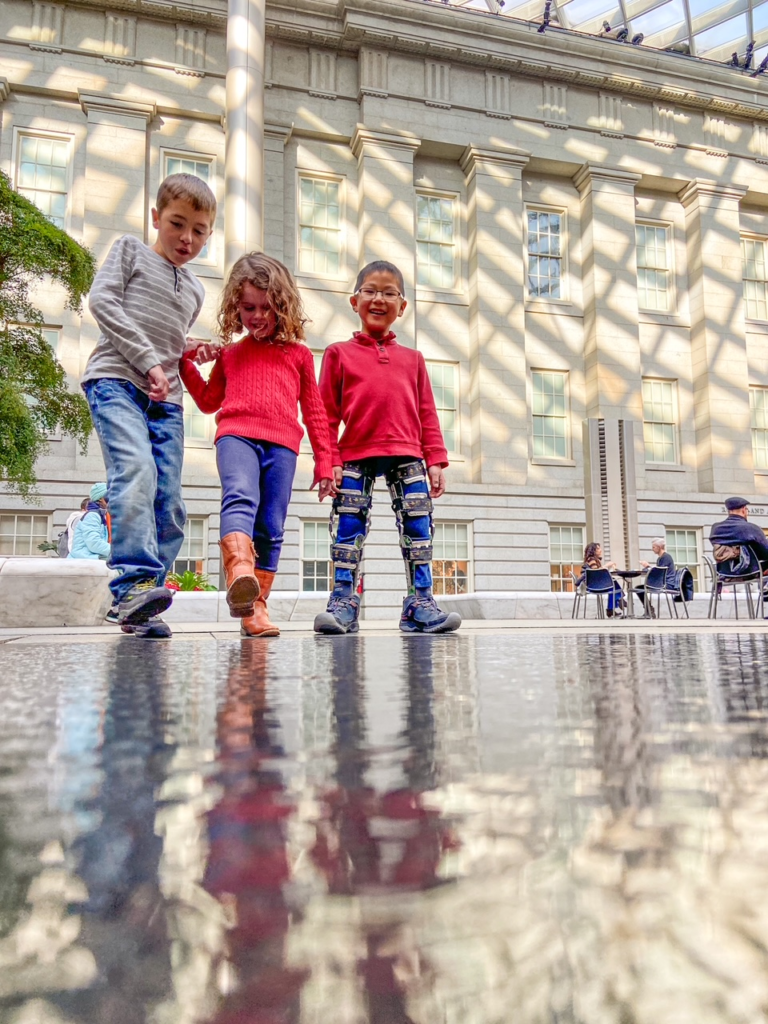 three kids playing with their reflections in the water below their feet. White columns in the background.