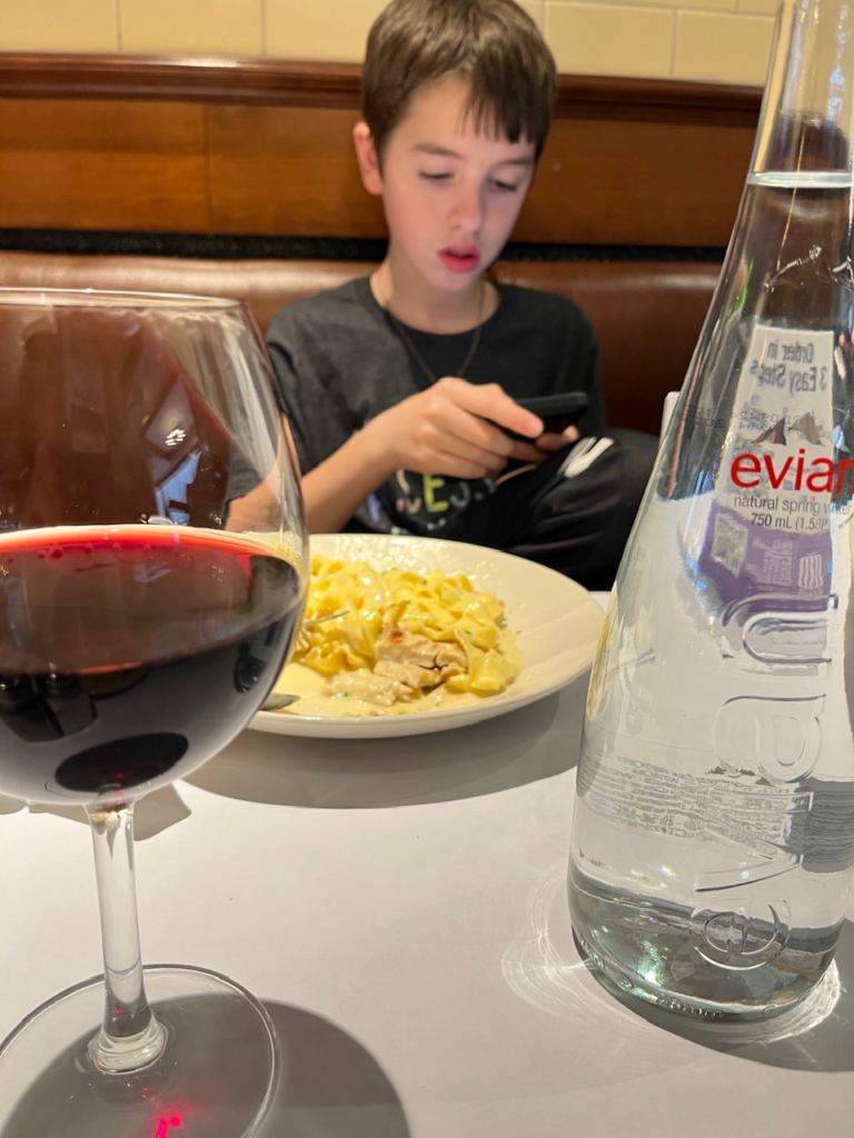 boy eating mac and cheese, glass of wine and bottle of Evian in foreground