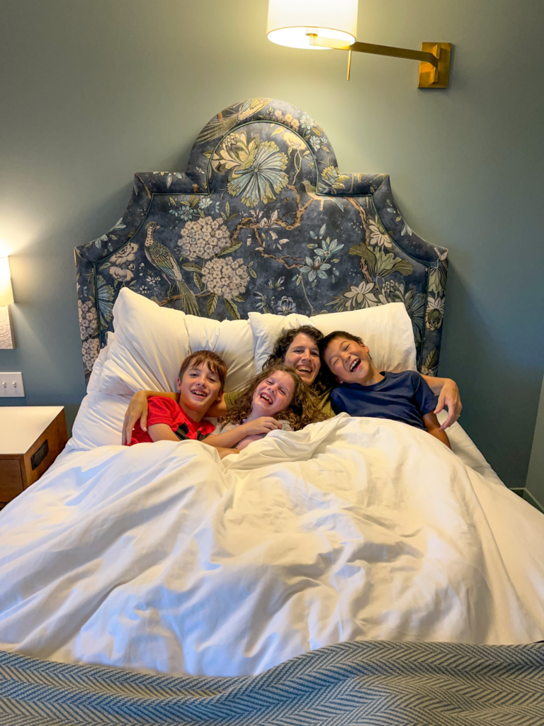 mom and three kids cuddling and laughing in hotel bed with big blue backboard.