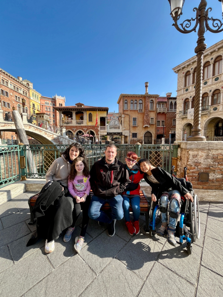 family, including little boy in wheelchair, older brother, younger sister, and mom and dad, sit in DisneySea's venice and smile at the camera. Arching bridge in the background.