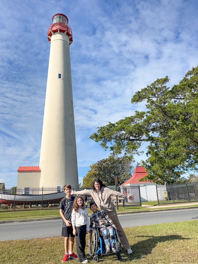 family - mom, little girl, and two little boys, one in a wheelchair - smiles in front of tall lighthouse with red cap