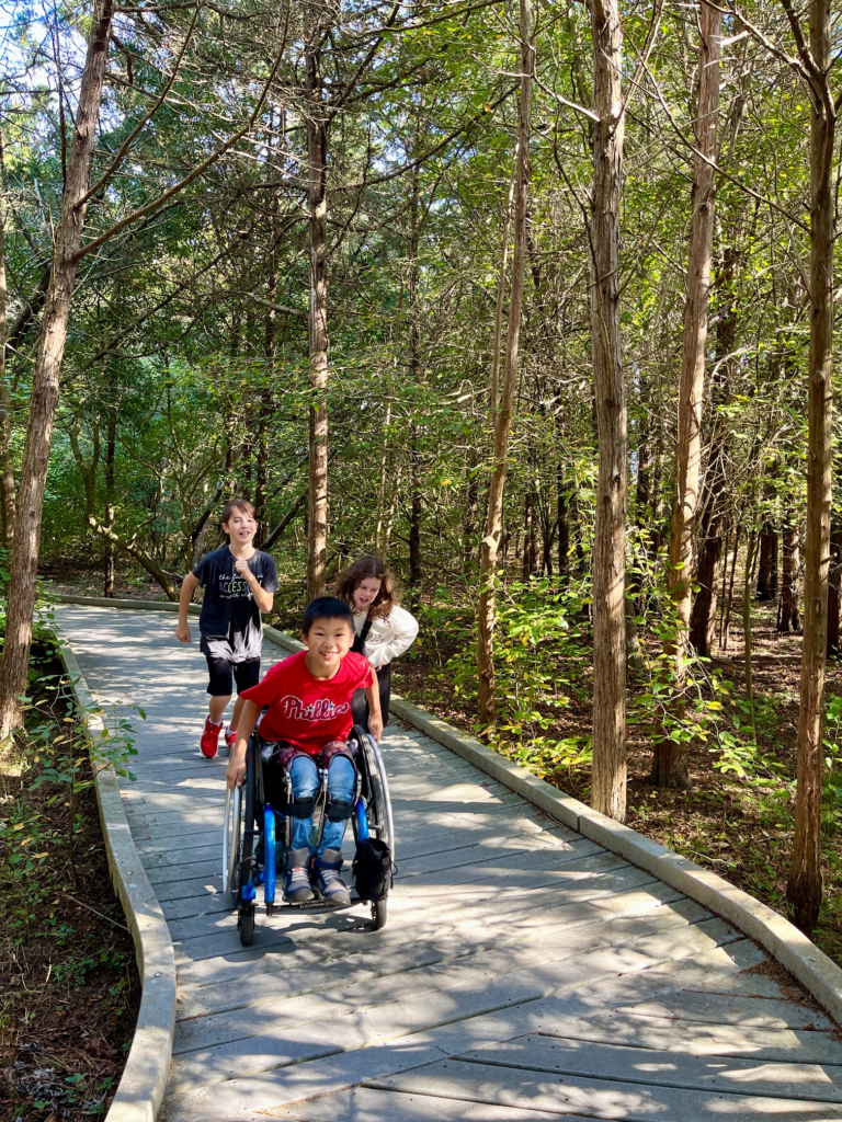 little boy in wheelchair leads the way down boardwalk path with sister and brother running after him