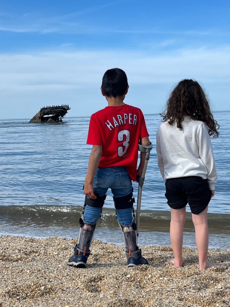 back of boy with forearm crutch and girl holding pants legs up facing the ocean with a sunken ship in the background
