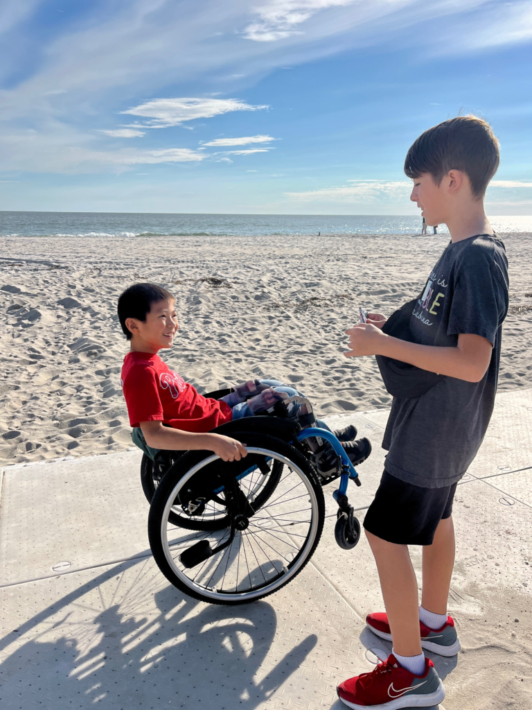 little bot doing a wheelie in his wheelchair on an accessible path through the sand, talking to his older brother. Ocean in the background