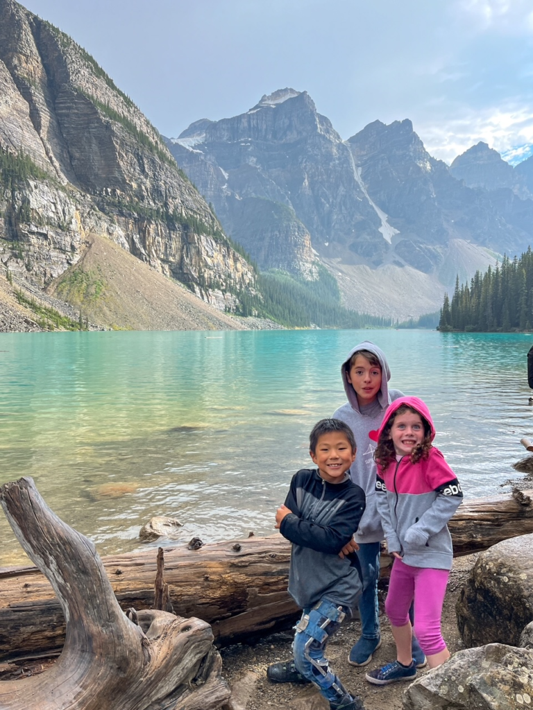 three children grin broadly in front of blue water and towering snow capped mountains