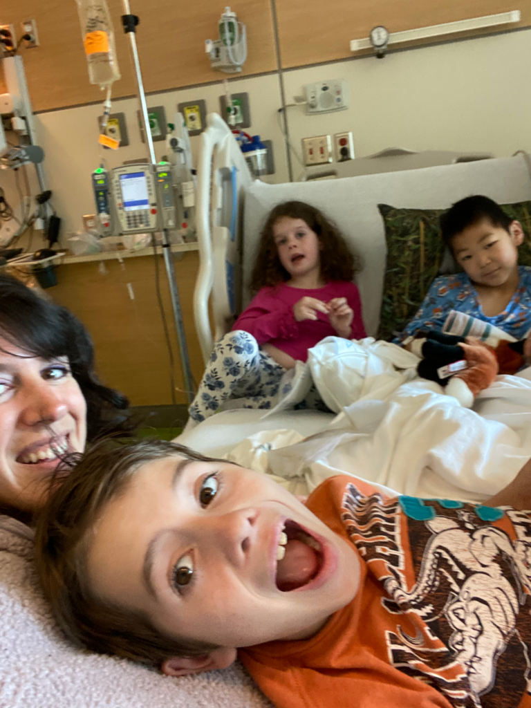 Young patient with sister, brother and mom in his hospital bed. All smile at camera. Big brother has his mouth open in a goofy yelling face.