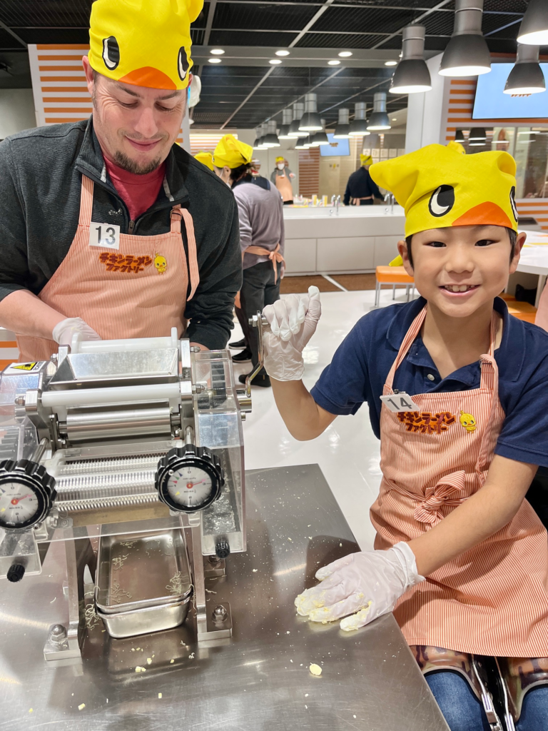 boy in wheelchair smiles at camera as he spins the handle on a noodle press