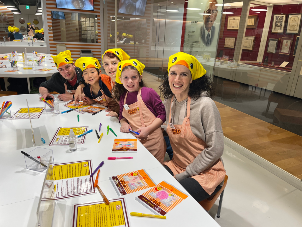 family in yellow duck bandanas and orange aprons smile at camera in Cup Noodle factory