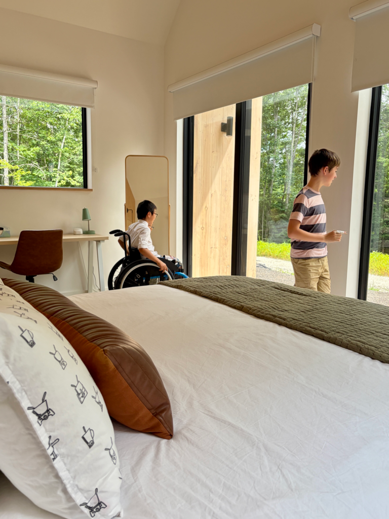 two boys look out large windows into the forest, white bed in foreground