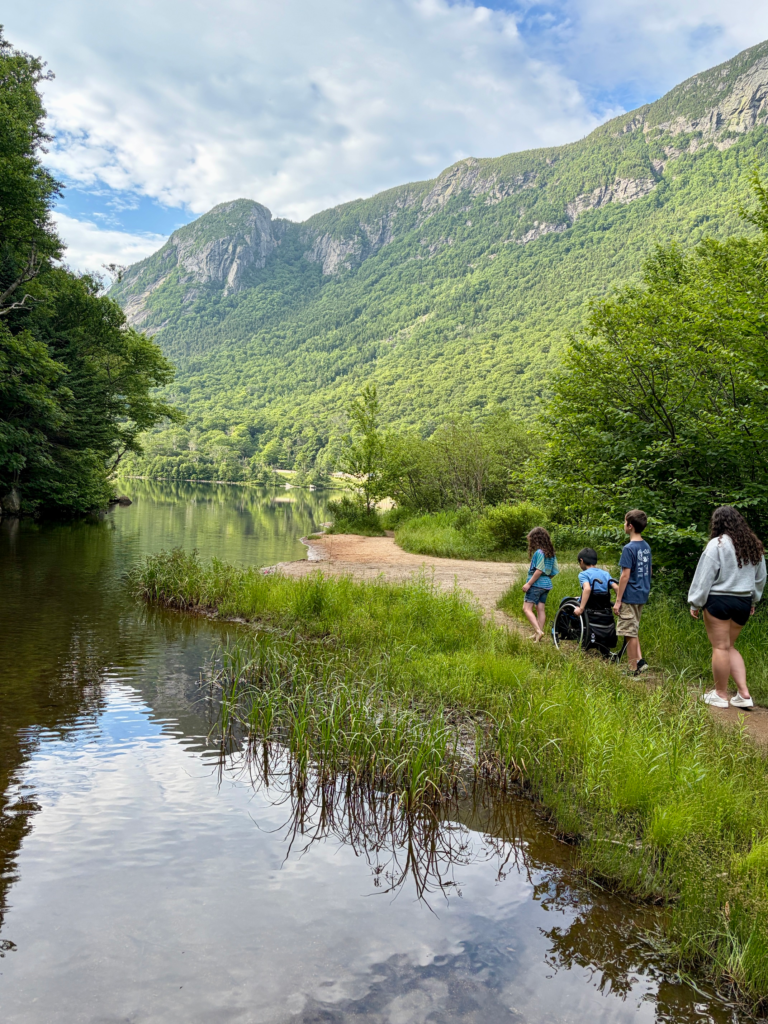 children walk/roll along edge of lake on sand trail with white mountains towering in the distance