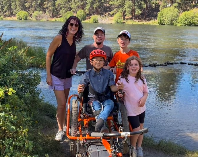 Mom, Dad, big brother, and little sister smile at camera from behind Jaden in the big orange AdvenChair. He's on a rugged trail with the river flowing behind