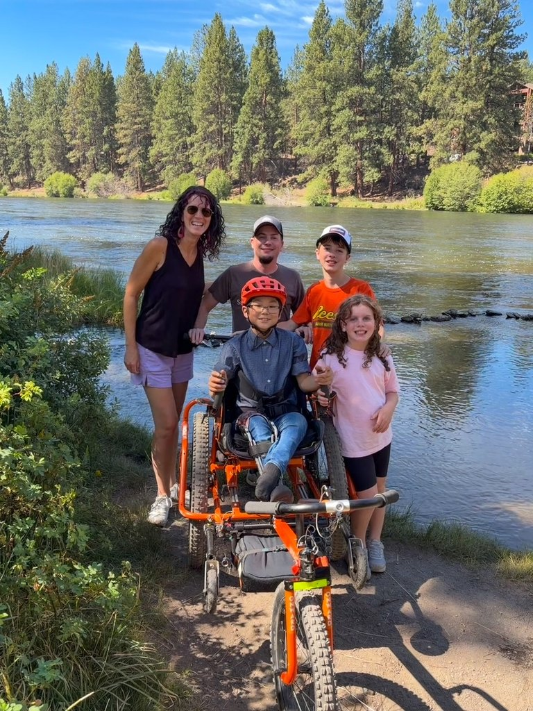 Mom, Dad, big brother, and little sister smile at camera from behind Jaden in the big orange AdvenChair. He's on a rugged trail with the river flowing behind.