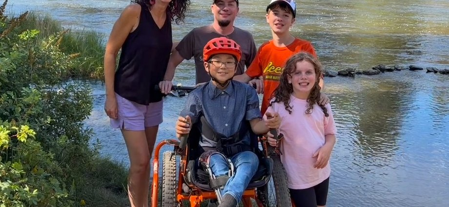 Mom, Dad, big brother, and little sister smile at camera from behind Jaden in the big orange AdvenChair. He's on a rugged trail with the river flowing behind